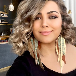 May include: A woman with long, wavy, light brown hair wearing large, long, feather earrings. The earrings are made of brown, beige, and teal feathers.