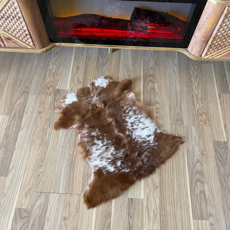 May include: A brown and white cowhide rug with a natural pattern. The rug is irregularly shaped and lies on a light-colored wooden floor. The rug is near a fireplace.