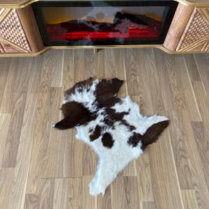 May include: A cowhide rug with a brown and white spotted pattern. The rug is irregularly shaped and lies on a wooden floor. The rug is in front of a wooden cabinet with a fireplace.