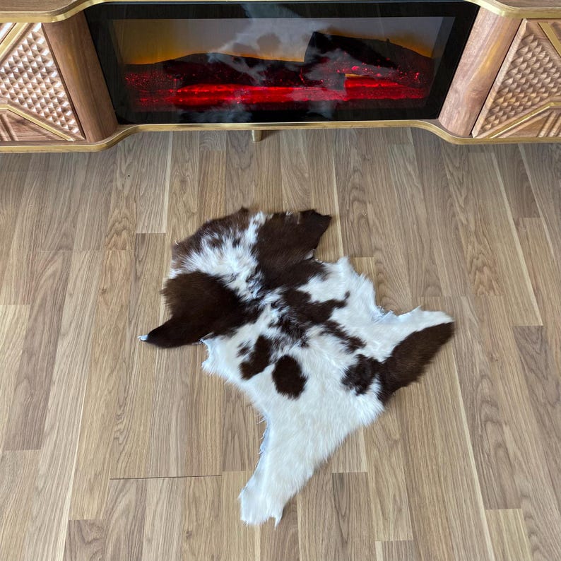 May include: A cowhide rug with a brown and white spotted pattern. The rug is irregularly shaped and lies on a wooden floor. The rug is in front of a wooden cabinet with a fireplace.
