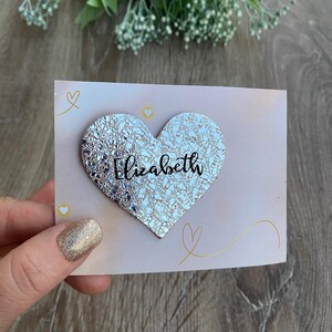 May include: A light pink card with a silver heart-shaped embellishment that reads "Elizabeth" in cursive. The card has gold heart and swirl accents. The background is a wooden surface.