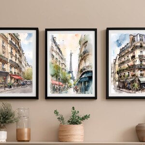 Puede incluir: Tres acuarelas enmarcadas que representan escenas callejeras parisinas. La pintura más a la izquierda muestra una calle bordeada de edificios, la pintura del medio muestra una calle con la Torre Eiffel en la distancia, y la pintura más a la derecha muestra un café con un edificio en el fondo.