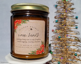 May include: A 7 oz beeswax candle in an amber glass jar with a gold lid. The label features the text Wildan Whimsy, warm hearth, stockings hung over a cozy fireplace, vanilla eggnog, roasting chestnuts, and handcrafted in Atlanta, with red poinsettia graphics.