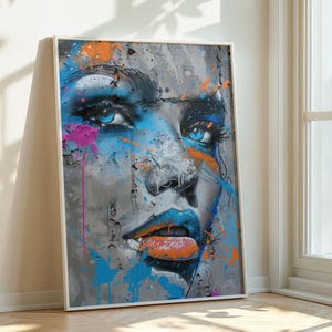 Puede incluir: Una impresión artística enmarcada que presenta un retrato estilizado del rostro de una mujer. La obra de arte utiliza una paleta de colores vibrantes de azul, naranja y rosa sobre un fondo gris, con efectos de salpicaduras de pintura. Los labios de la mujer son de un azul brillante con una lengua naranja.