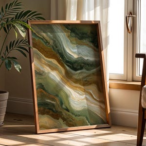 May include: A framed abstract art print with a flowing design in shades of green, gold, and white. The artwork is displayed in a wooden frame and leans against a wall near a window, showcasing a modern aesthetic.