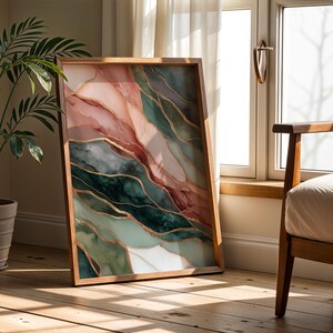 May include: A framed abstract art print featuring a watercolor design with shades of pink, green, and gold. The artwork is in a wooden frame and leans against a wall near a window.