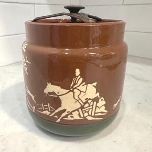 May include: Brown ceramic jar with a lid, featuring a cream-colored equestrian scene. The jar has a green base and a dark metal lid. The design includes a rider on a horse jumping over a fence. A decorative kitchen storage container.