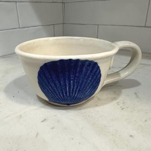 Puede incluir: Taza de cerámica color crema con un gran diseño de concha azul. La taza tiene forma redondeada, un asa robusta y una abertura ancha. La concha está detallada con finas líneas, añadiendo textura. Una pieza sencilla y elegante para disfrutar de bebidas.