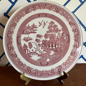 Peut inclure: Une grande assiette en céramique blanche avec un motif de saule rose. L'assiette présente une scène de paysage chinois traditionnelle avec une pagode, des arbres et un pont. L'assiette est sur un support en métal doré.
