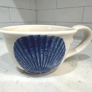 Puede incluir: Taza de cerámica color crema con un gran diseño de concha azul. La taza tiene forma redondeada, un asa curva y una abertura ancha. El diseño de concha está centrado en la parte delantera.