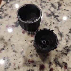 May include: Two black, cylindrical plastic components, possibly parts of a larger mechanism, are displayed. One component is a cup-shaped piece with a ridged exterior. The other is a smaller, circular piece with internal structures.