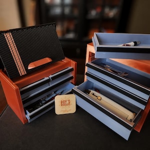 May include: Two miniature orange and light gray storage chests with multiple drawers. The left chest features a black carbon fiber patterned top with three copper stripes. A square tan card with the text ROY'S WONDERS is placed between them.