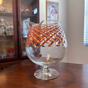 May include: A clear glass pitcher with a gold and red geometric pattern around the middle. The pitcher has a round body and a short stem.