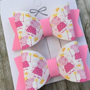 May include: Two pink and white hair bows with a cartoon pig wearing a crown and wings pattern.