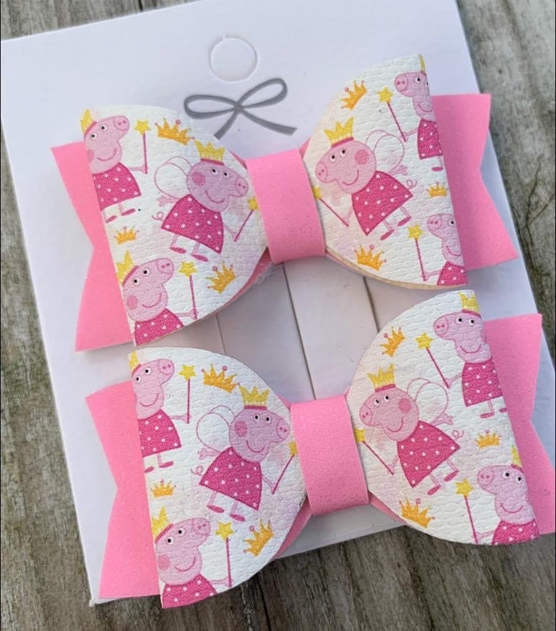 May include: Two pink and white hair bows with a cartoon pig wearing a crown and wings pattern.