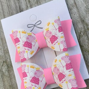 May include: Two pink hair bows with a white fabric featuring a repeating pattern of a cartoon pig wearing a crown and fairy wings. The bows are tied together with a pink ribbon.