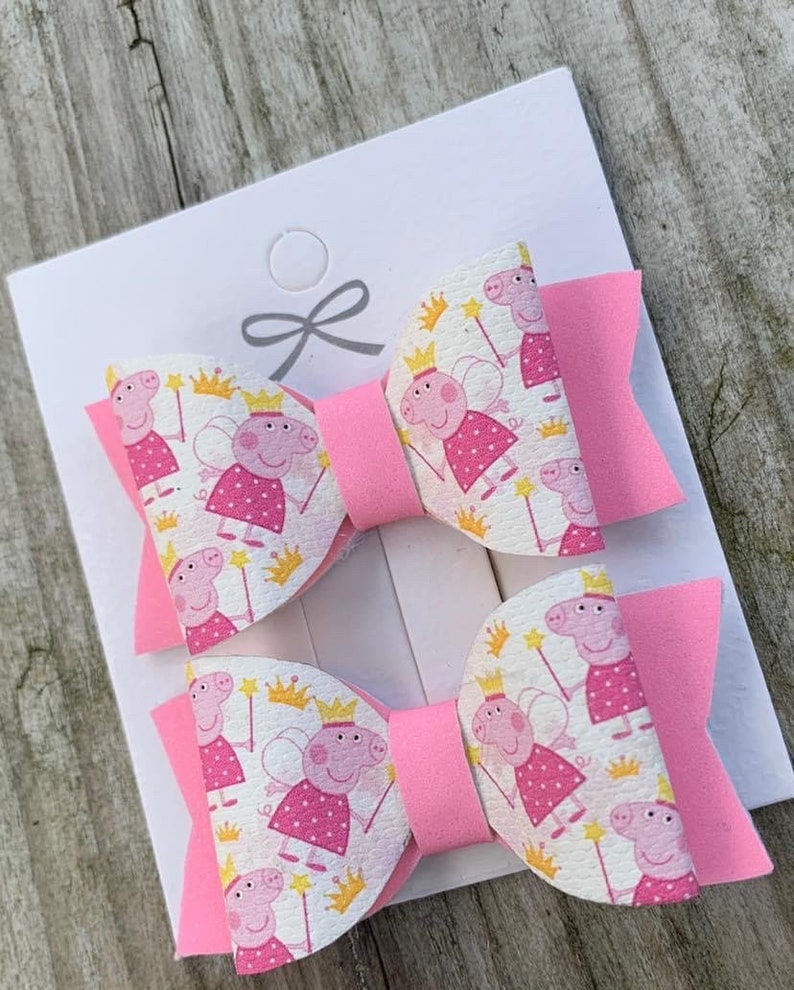 May include: Two pink hair bows with a white fabric featuring a repeating pattern of a cartoon pig wearing a crown and fairy wings. The bows are tied together with a pink ribbon.
