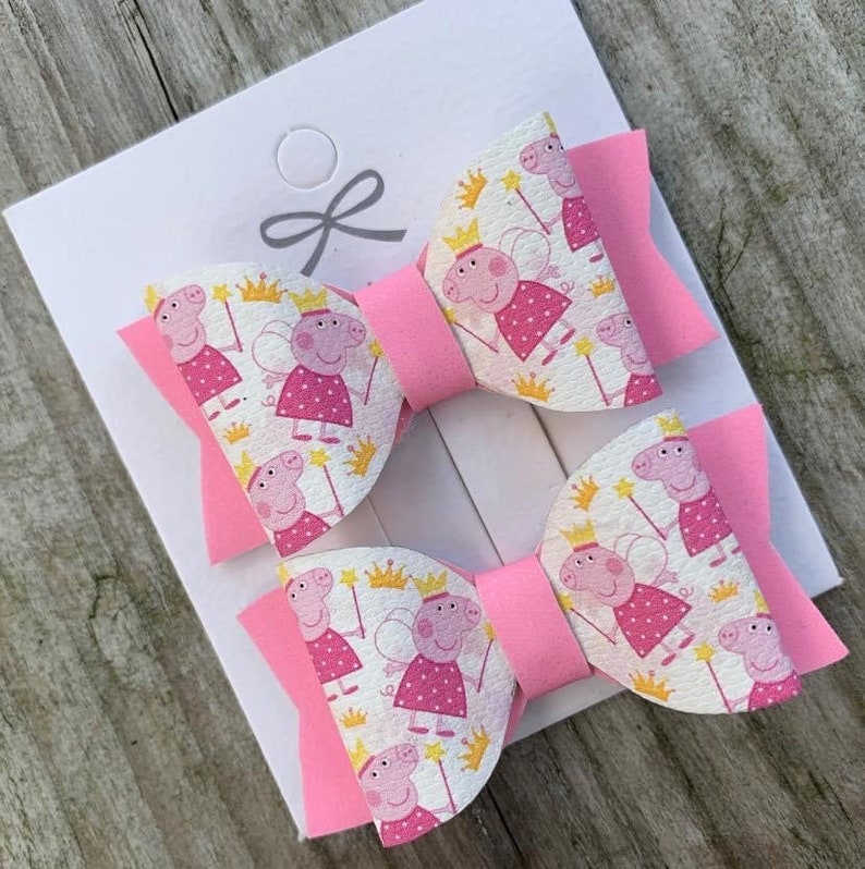 May include: Two pink hair bows with a white background featuring a repeating pattern of pink pigs wearing crowns and fairy wings. The bows are made of felt and have a pink center.