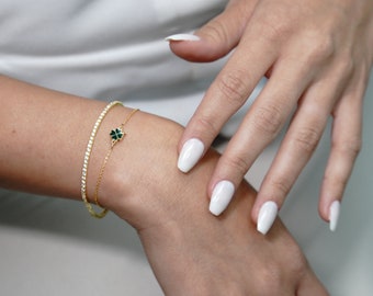 May include: Two gold bracelets on a wrist. One is a tennis bracelet with small, clear stones. The other is a delicate chain with a four-leaf clover charm with green enamel. The background is a white shirt.