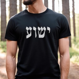 May include: A black short-sleeved t-shirt featuring the Hebrew word Yeshua printed in large, light gray lettering across the chest. The garment has a standard crew neckline and a straight hemline, providing a simple and clean design.