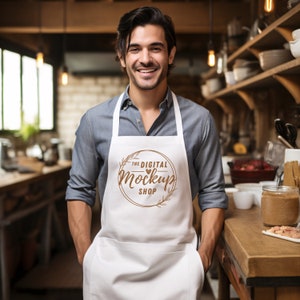 May include: A man wearing a white apron with a brown logo that says "The Digital Mockup Shop" with a heart symbol. He is standing in a kitchen with a wooden countertop and shelves.
