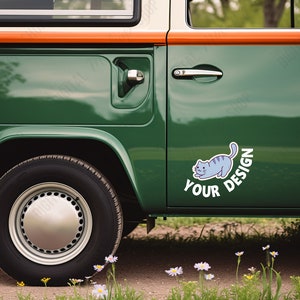 May include: A green and white vintage van with a sticker of a blue cat and the words "YOUR DESIGN". The van has a silver hubcap and a black tire. The van is parked on a dirt road with wildflowers.