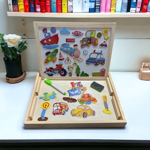 May include: A wooden magnetic board with a white dry-erase surface and a separate play area. The board features colorful magnetic pieces of vehicles, traffic signs, and other objects. The play area has a green marker and a black eraser.