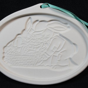 May include: White ceramic cookie cutter shaped like a bunny holding a basket of eggs. The cutter has a hole at the top for hanging.