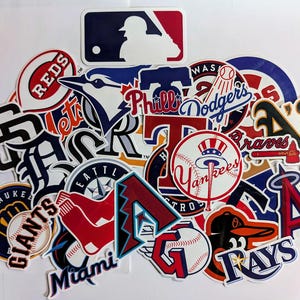 Könnte beinhalten: Eine Sammlung farbenfroher Baseball-Team-Logo-Aufkleber, darunter die Yankees, Dodgers und Braves. Die Aufkleber zeigen verschiedene Formen und Designs mit Teamnamen und Logos. Das MLB-Logo ist ebenfalls vorhanden.