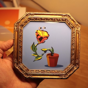 May include: An octagonal gold-toned frame containing a print of a stylised flower with yellow and red petals, green leaves, and two eyes on stalks, growing out of a brown terracotta-coloured pot against a solid light blue background.