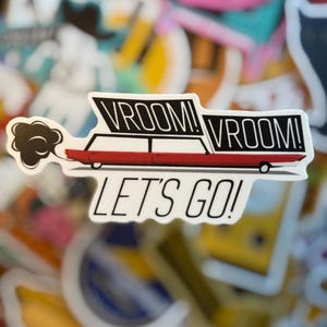 May include: A sticker featuring a red car with the words "VROOM! VROOM!" in black and white above it. Below the car, the text "LET'S GO!" is displayed. A cloud of black smoke emanates from the car.