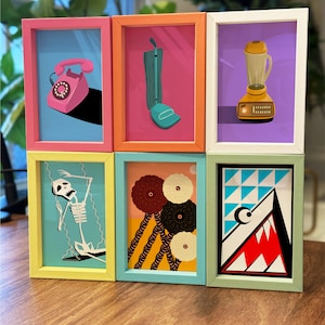 May include: Six framed art prints with colorful illustrations. The prints feature a pink rotary phone, a green vacuum cleaner, a yellow blender, a skeleton on a swing, abstract flowers, and a geometric shark design. Each print is in a different colored frame.