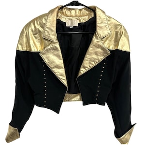 Puede incluir: Chaqueta corta negra y dorada. La chaqueta tiene cuello y puños dorados metálicos, con cuerpo negro. Pequeños tachuelas doradas bordean el frente. La chaqueta está colgada de una percha negra. Diseño llamativo.
