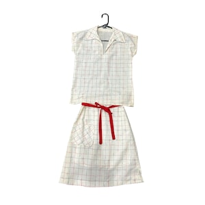 May include: A vintage two-piece outfit featuring a collared, short-sleeved top and a midi skirt. The cream-colored fabric has a grid pattern with red and blue lines. A red belt cinches the skirt's waist, and the outfit hangs on a black hanger.