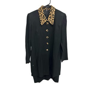 Puede incluir: Vestido negro con cuello estampado de leopardo y botones dorados. El vestido está colgado de una percha negra. El vestido tiene mangas largas y un corte entallado. Los botones son redondos y tienen un acabado dorado.