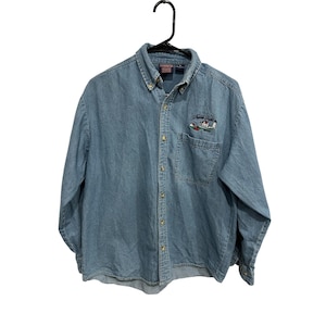 May include: Light blue denim button-down shirt with long sleeves. The shirt has a small embroidered design on the chest pocket with the words "Middle Bay" and a small graphic. The shirt is hanging on a black hanger.