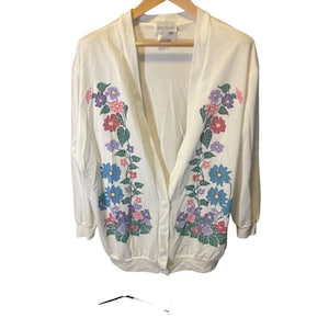 May include: A white long-sleeve cardigan featuring a V-neckline and a button-front closure. The garment is decorated with two symmetrical floral patterns in shades of blue, purple, pink, and green, extending from the shoulders to the hem.