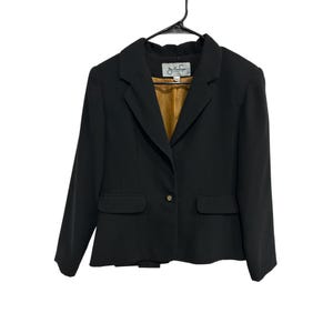 May include: A black blazer featuring a notched lapel, long sleeves, and two front flap pockets. The jacket has a single button closure at the center and a contrasting mustard yellow interior lining. A label with the text Julian Taylor is visible.