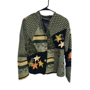 Puede incluir: Una chaqueta de manga larga con un diseño de patchwork que presenta varios paneles de tela en verde, negro y dorado. La prenda incluye motivos de estrellas bordados en amarillo y naranja, un escote en V y tres botones circulares al frente.