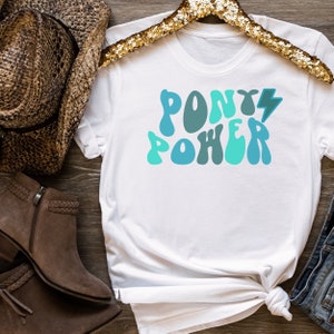 Può includere: Maglietta bianca con una grafica blu e verde che dice "Pony Power" con un fulmine.