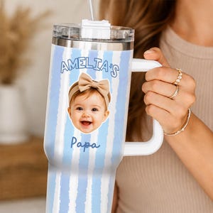 K&ouml;nnte beinhalten: Ein personalisierter Becher mit wei&szlig;em Griff und transparentem Deckel. Der Becher hat ein blau-wei&szlig; gestreiftes Muster und ein Foto eines Babys mit Schleife. Der Text "AMELIA'S" und "Papa" sind ebenfalls sichtbar.
