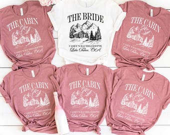 Peut inclure: Plusieurs t-shirts blancs et mauves. Un t-shirt blanc avec l'inscription "THE BRIDE" et un motif de cabane. Les t-shirts mauves indiquent "THE CABIN Crew" avec un motif de cabane. Tous les t-shirts portent l'inscription "Casey's Bachelorette Lake Tahoe, CA".