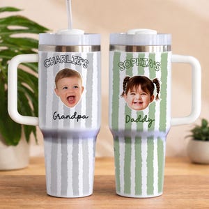 K&ouml;nnte beinhalten: Zwei personalisierte Reisebecher mit wei&szlig;en Griffen und Deckeln. Ein Becher hat ein grau-wei&szlig;es Streifenmuster mit einem Babyfoto und dem Text "Charlie's Grandpa". Der andere Becher hat ein gr&uuml;n-wei&szlig;es Streifenmuster mit einem Babyfoto und dem Text "Sophia's Daddy".