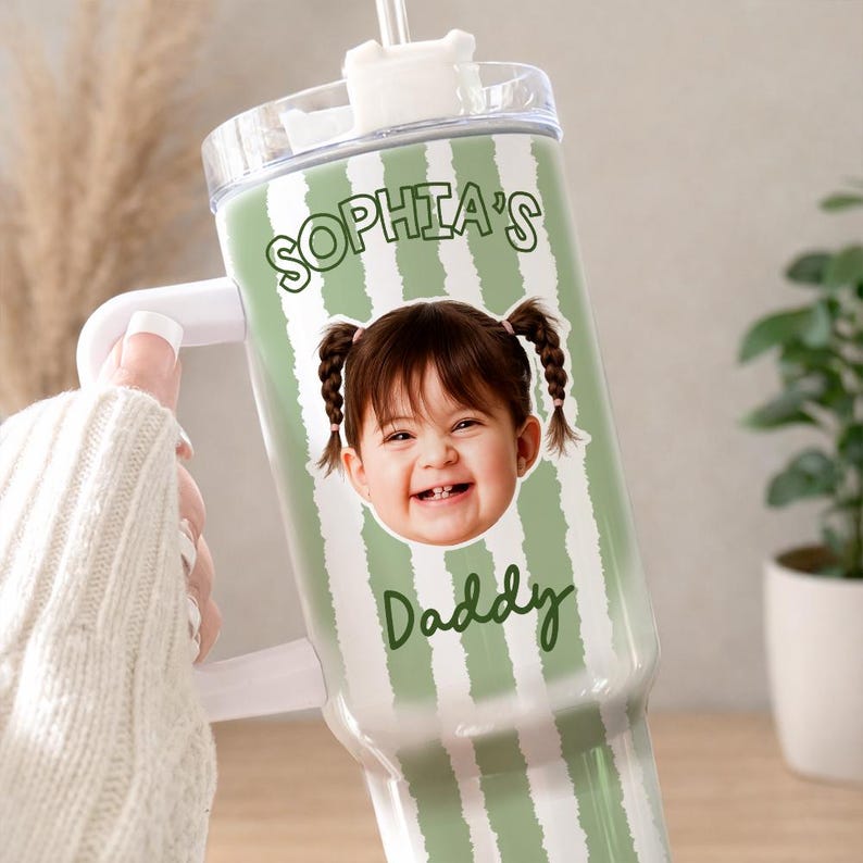 K&ouml;nnte beinhalten: Ein personalisierter Becher mit wei&szlig;em Griff und transparentem Deckel. Der Becher hat ein gr&uuml;n-wei&szlig; gestreiftes Design mit dem Namen "Sophia's" und dem Wort "Daddy" in gr&uuml;ner Schrift. Ein Foto eines l&auml;chelnden Kindes ist ebenfalls auf dem Becher abgebildet.