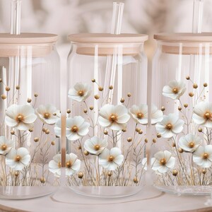 Puede incluir: Tres tarros de vidrio transparente con tapas de madera y dise&ntilde;os florales blancos. Cada tarro tiene una pajita que sobresale de la tapa.