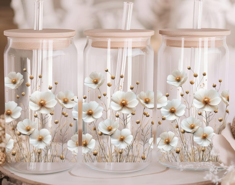 Puede incluir: Tres tarros de vidrio transparente con tapas de madera y dise&ntilde;os florales blancos. Cada tarro tiene una pajita que sobresale de la tapa.