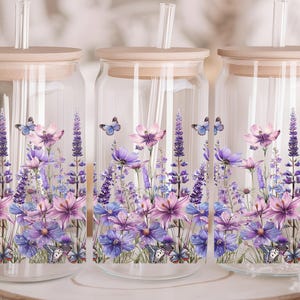 Puede incluir: Tres tarros de vidrio con tapas de madera y pajitas. Cada tarro tiene un dise&ntilde;o floral con flores y mariposas moradas, rosas y azules. Los tarros son perfectos para guardar bebidas u otros art&iacute;culos.