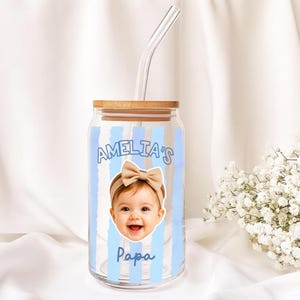 K&ouml;nnte beinhalten: Ein durchsichtiges Glas im Dosen-Design mit Bambusdeckel und gebogenem Glasstrohhalm. Der Becher zeigt ein Babyfoto mit Schleife, den Namen "AMELIA'S" und das Wort "Papa" in Blau, mit blauen vertikalen Streifen.