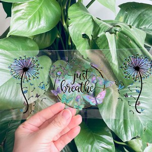 May include: Clear sticker featuring a dandelion design with the text "just breathe" in a gold circle. The dandelion is iridescent and has blue and purple hues. There are butterflies around the circle.