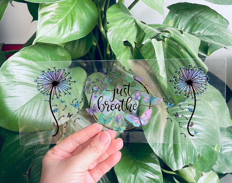May include: Clear sticker featuring a dandelion design with the text "just breathe" in a gold circle. The dandelion is iridescent and has blue and purple hues. There are butterflies around the circle.
